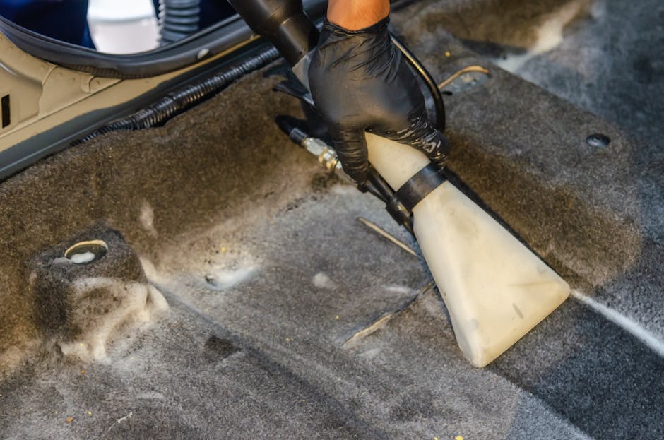 A person wearing a black glove is using a handheld steam cleaning machine to deep clean the grey fabric car upholstery. The device emits a concentrated stream of steam onto the surface, removing dirt and stains, with visible foam and moisture indicating active cleaning. Surrounding the section of the upholstery are the edges of a vehicle's interior, showing parts of the floor and side panels. The setting is well-lit, highlighting the cleanliness process as part of surface cleaning and maintenance, consistent with Kingston Carpet Cleaning's same-day upholstery cleaning service at the Bentall Centre in Kingston.