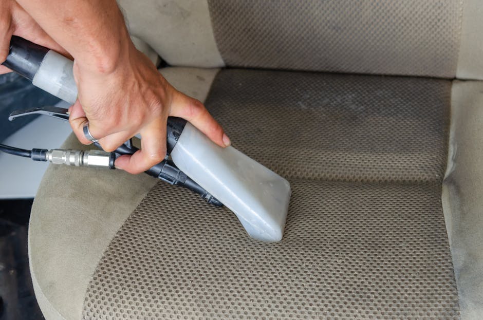 A person wearing a black glove is using a handheld steam cleaning machine to deep clean the grey fabric car upholstery. The device emits a concentrated stream of steam onto the surface, removing dirt and stains, with visible foam and moisture indicating active cleaning. Surrounding the section of the upholstery are the edges of a vehicle's interior, showing parts of the floor and side panels. The setting is well-lit, highlighting the cleanliness process as part of surface cleaning and maintenance, consistent with Kingston Carpet Cleaning's same-day upholstery cleaning service at the Bentall Centre in Kingston.
