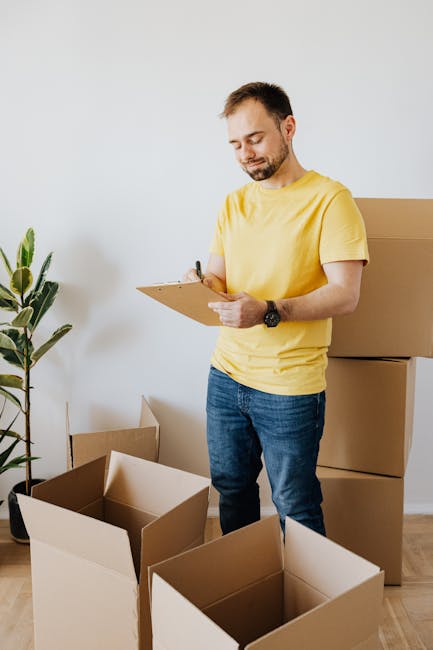 A man with a beard wearing a yellow t-shirt and jeans is standing in a bright room with white walls, surrounded by open cardboard boxes on the wooden floor. He is holding a clipboard and a pen, appearing to be inspecting or recording items, with a large potted plant with green leaves nearby. This scene depicts a residential setting related to move-in or move-out processes, emphasizing thorough surface cleaning and organisation, as part of end of tenancy cleaning services offered by Kingston Carpet Cleaning. The room is well-lit with natural light, and the environment is clean and uncluttered, highlighting the importance of hygiene and proper preparation during property handovers.