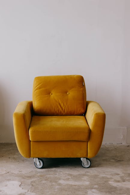 A mustard yellow upholstered armchair with a cushioned backrest and armrests, placed on a concrete floor against a plain white wall. The chair has casters attached to its base, allowing for mobility.