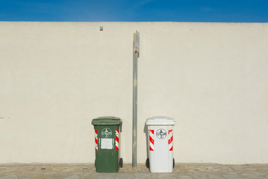 Three residential wheelie bins are positioned on a paved curb side in front of a dense green hedge. The black bin on the left is slightly smaller, with a closed lid and a set of wheels at the base. The middle bin is larger, blue, with a closed lid, and has two wheels visible. The green bin on the right is of similar size to the blue one, with a top lid closed, and no visible wheels. The surface beneath the bins is a smooth asphalt, with a concrete curb separating the pavement from the grassy area. Bright daylight illuminates the scene, highlighting the clean surfaces of the bins and the lush appearance of the hedge. This setting illustrates typical domestic waste disposal, and the clean, well-maintained appearance of the bins aligns with professional cleaning standards, as promoted by Kingston Carpet Cleaning, in relation to Kingston council rules for waste and cleaning disposal.