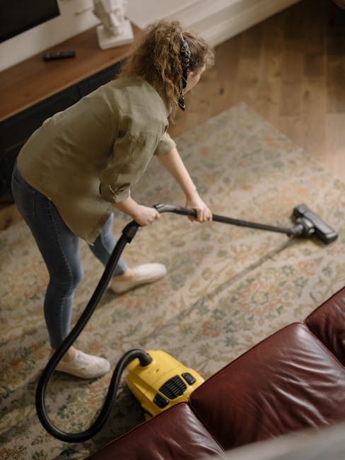 A woman with curly hair, wearing a beige jacket, dark jeans, and white shoes, is performing surface cleaning on a large, patterned area rug in a living room. She is using a yellow vacuum cleaner with a hose attachment to deep clean the rug, which features intricate floral designs in muted colors. The room has wooden flooring partially visible around the rug, with soft, warm lighting illuminating the space. In the background, a wooden sideboard with decorative items and a television can be seen, contributing to a cozy, tidy environment. This image demonstrates domestic cleaning practices that Kingston Carpet Cleaning offers for maintaining hygiene and cleanliness of carpets and rugs in residential settings.