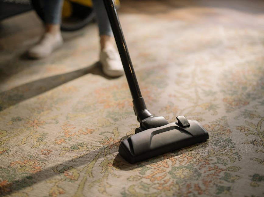 Close-up of a vacuum cleaner head actively cleaning a patterned, beige and green residential carpet in a well-lit room. The vacuum's black floor attachment is pressed flat against the carpet, with a visible roller brush at the base. In the background, a person's legs are partially visible, standing nearby, wearing casual footwear. The carpet surface appears free of dust and debris, indicating thorough deep cleaning. The scene emphasizes surface cleaning and maintenance as part of professional domestic cleaning services offered by Kingston Carpet Cleaning, aligning with the Kingston KT1 area guide page.