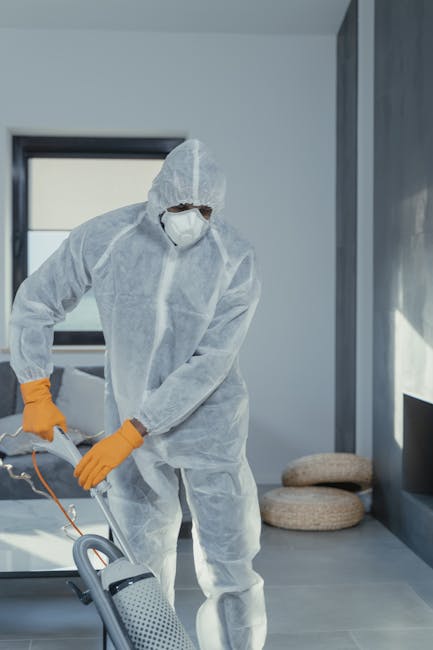 A professional cleaning specialist dressed in a full white protective suit, face mask, and orange gloves, operating a vacuum cleaner in a modern, well-lit residential space. The room features large windows allowing natural light, with a calm grey color palette on the walls and floor. In the background, two round, woven cushions are placed on the floor beside a dark grey sofa. This scene depicts surface cleaning and deep cleaning activities aimed at maintaining hygiene and pristine condition of the living environment. Kingston Carpet Cleaning offers comprehensive domestic cleaning and surface sanitisation services to ensure a spotless and healthy home, as highlighted in their Kingston cost guide.