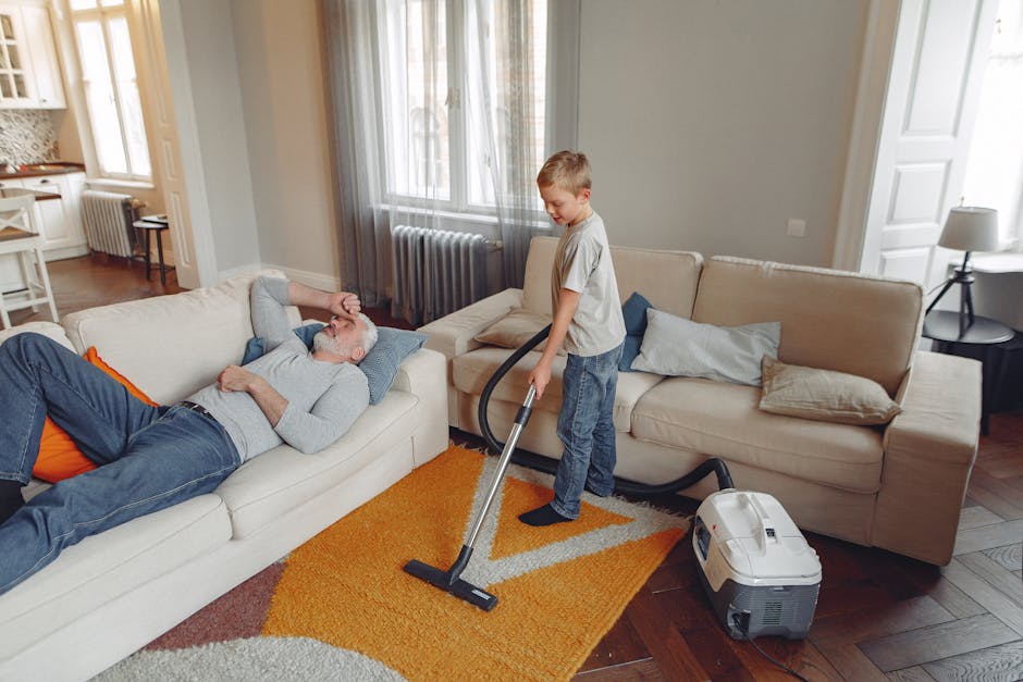 A young boy is vacuuming a beige fabric sofa with a handheld vacuum cleaner, while an older man with gray hair and a beard is lying on the sofa, resting with his eyes closed and hands behind his head. The room features wooden flooring, a bright orange and beige area rug, and large windows with sheer curtains allowing natural light to illuminate the space. The sofa is accompanied by light-colored cushions, and nearby there is a small black side table with a white lamp. The overall scene depicts domestic surface cleaning, emphasizing effective deep cleaning and sanitisation practices, as seen in the neat, dust-free environment with clean surfaces, consistent with professional cleaning standards offered by Kingston Carpet Cleaning.