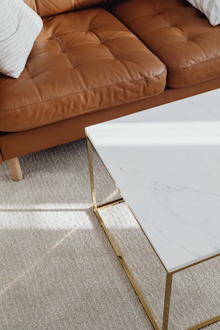 A close-up view of a brown leather sofa in a living room, featuring tufted cushions and white decorative pillows. Adjacent to the sofa is a white marble-top coffee table with gold metal legs. The area is illuminated by natural light, highlighting the clean and polished appearance of the furniture. The room has a beige textured carpet that appears well-maintained and free of dust or debris. This setting illustrates a neat and hygienic domestic space, showcasing surface cleaning and deep cleaning practices for upholstery and furnishings, with services provided by Kingston Carpet Cleaning. The overall scene emphasizes clarity and freshness in a contemporary living room environment.