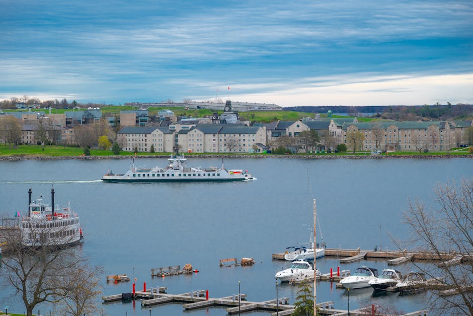 A scenic view of the Kingston riverside featuring a calm river with several moored boats, including a large paddle steamer with black chimneys, surrounded by a walkway and docks. Across the river, the