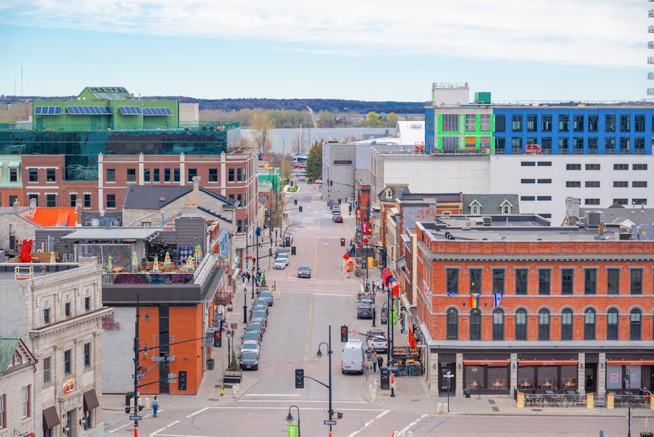 An aerial view of a city street in Kingston featuring a mix of historic and modern buildings. The foreground shows brick and stone structures with large windows, some with flags and decorative elements. The street is lined with parked cars, pedestrians, and street lamps, with a clear road surface. In the background, there are taller contemporary buildings with glass and steel facades, including a distinctive green rooftop structure on one building. A river can be seen in the distance beyond the cityscape, under a partly cloudy sky, indicating a typical urban scene in Kingston. This image emphasizes the vibrant mix of architecture and active street life, relevant to Kingston Carpet Cleaning’s focus on maintaining cleanliness in diverse urban environments.