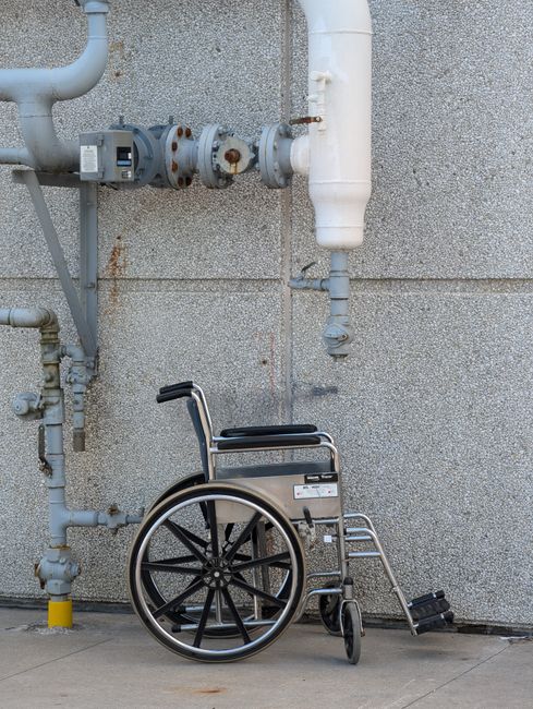 A silver wheelchair with black armrests and large spoked wheels is positioned on a concrete pavement against a grey textured exterior wall. Visible pipes and valves run horizontally and vertically along the wall above the wheelchair, some showing signs of rust and corrosion. The wheeled mobility aid is clean and appears ready for use, situated near a corner of the building, with natural lighting casting soft shadows on the surface. The image emphasizes accessible infrastructure in a commercial or public setting, relevant to maintenance and access considerations for Kingston Carpet Cleaning's first aid or accessibility-related services.